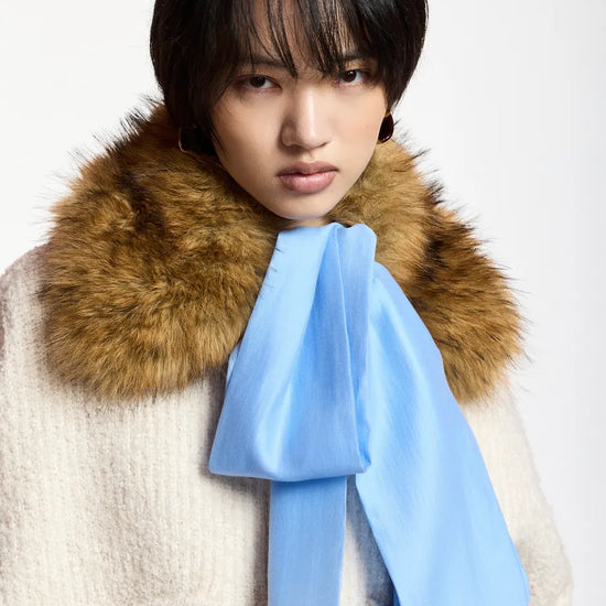A model wearing a cream-colored textured coat with a large brown fur collar and a light blue satin bow tied around the neck, set against a plain white background.