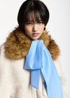 A model wearing a cream-colored textured coat with a large brown fur collar and a light blue satin bow tied around the neck, set against a plain white background.