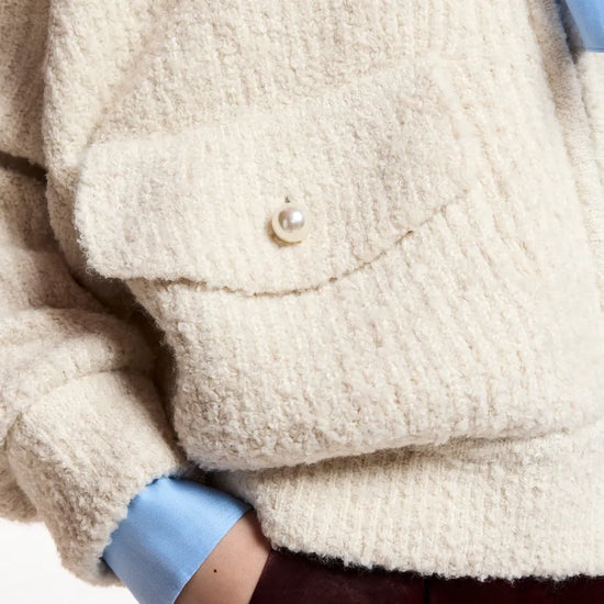Close-up of a person wearing a chunky cream bouclé cardigan with a pearl button, layered over a light blue collared shirt and dark burgundy trousers.