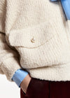 Close-up of a person wearing a chunky cream bouclé cardigan with a pearl button, layered over a light blue collared shirt and dark burgundy trousers.