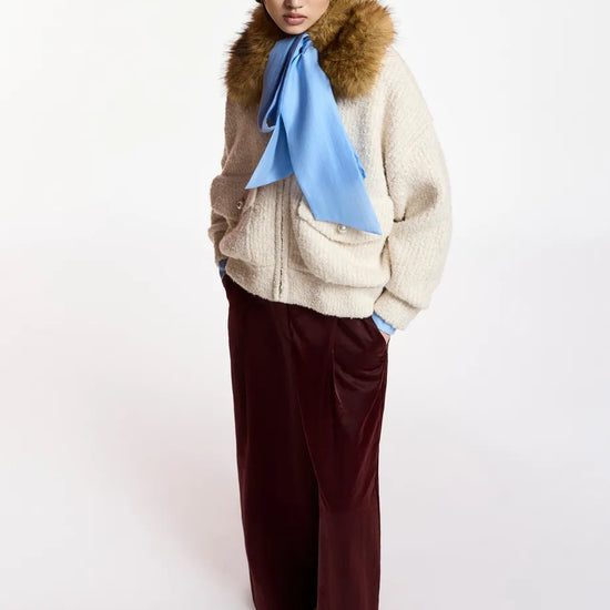 A model wearing a cream-colored knit zip-up jacket with a brown fur-trimmed collar, a light blue silk scarf, and dark burgundy wide-leg trousers, photographed against a plain white studio background.