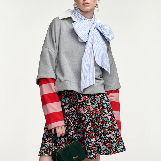 A model wearing a gray cropped sweatshirt with red and white striped sleeves, a blue and white striped bow tie scarf, a red and white floral mini skirt, and holding a dark green handbag against a light gray background.