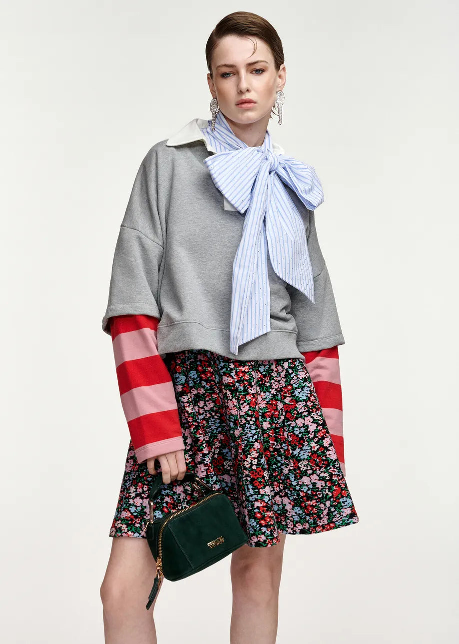 A model wearing a gray cropped sweatshirt with red and white striped sleeves, a blue and white striped bow tie scarf, a red and white floral mini skirt, and holding a dark green handbag against a light gray background.