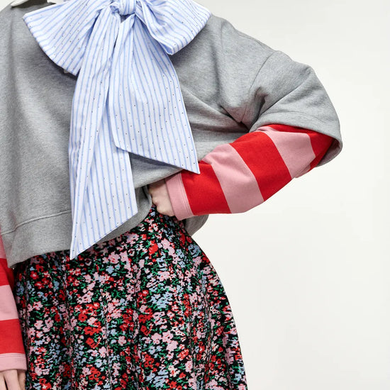 A model wearing a gray cropped sweatshirt with red and pink striped sleeves, a light blue and white striped bow tie, and a red, pink, and green floral skirt.