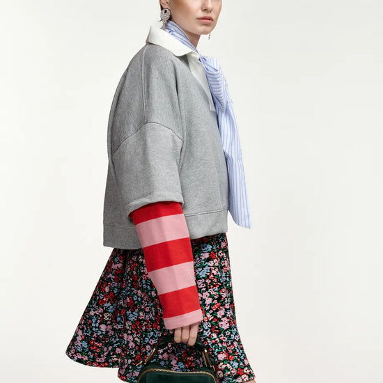 A model is shown from a side-back angle wearing a gray cropped jacket with red and pink striped knit sleeves, a blue and white striped shirt, a floral skirt, and holding a dark green handbag against a plain light background.
