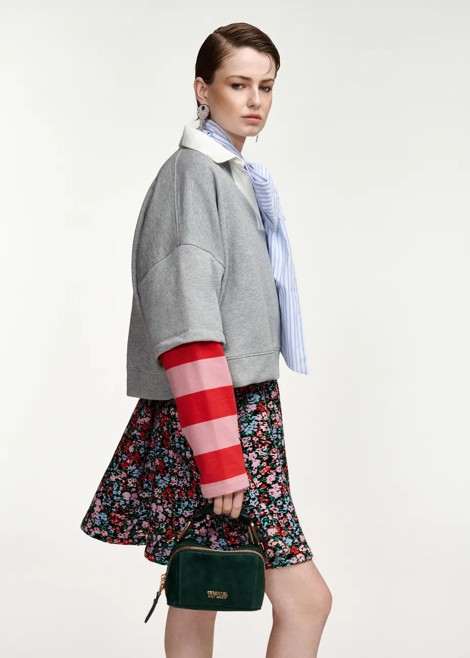 A model is shown from a side-back angle wearing a gray cropped jacket with red and pink striped knit sleeves, a blue and white striped shirt, a floral skirt, and holding a dark green handbag against a plain light background.