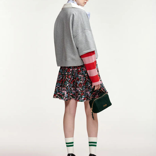 A model is shown from the back wearing a gray cropped cardigan with red and white striped sleeves, a floral mini skirt, green and white striped socks, and black ankle boots, while holding a dark green handbag against a light gray studio background.