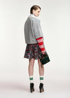 A model is shown from the back wearing a gray cropped cardigan with red and white striped sleeves, a floral mini skirt, green and white striped socks, and black ankle boots, while holding a dark green handbag against a light gray studio background.
