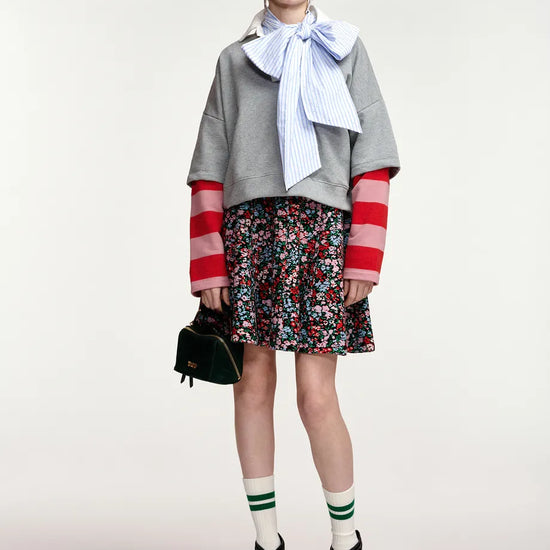 A model stands against a light gray studio background wearing a gray cropped sweater with red-and-white striped sleeves, a light blue striped bow tie, a dark floral mini skirt, green-and-white striped socks, black-and-white heeled shoes, and holding a small black handbag.