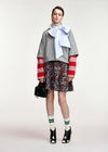A model stands against a light gray studio background wearing a gray cropped sweater with red-and-white striped sleeves, a light blue striped bow tie, a dark floral mini skirt, green-and-white striped socks, black-and-white heeled shoes, and holding a small black handbag.