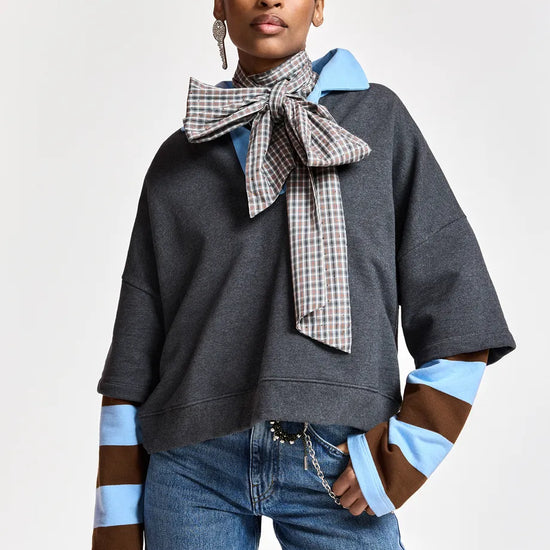 A model wearing a dark gray oversized sweatshirt with light blue and brown striped cuffs, a large checkered bow tie, and blue jeans, photographed against a light gray studio background.