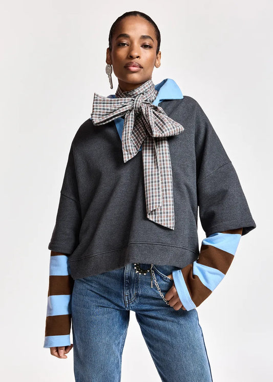 A model wearing a dark gray oversized sweatshirt with light blue and brown striped cuffs, a large checkered bow tie, and blue jeans, photographed against a light gray studio background.