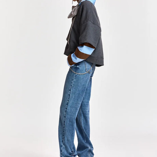 A model is shown in profile wearing a dark gray cropped jacket with light blue and brown striped cuffs, blue jeans, and black pointed shoes, against a plain light gray studio background.