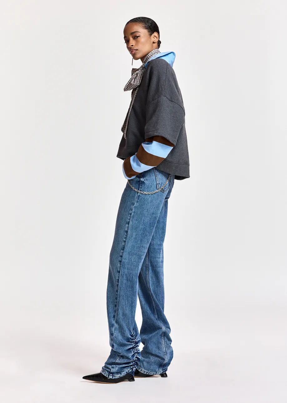 A model is shown in profile wearing a dark gray cropped jacket with light blue and brown striped cuffs, blue jeans, and black pointed shoes, against a plain light gray studio background.