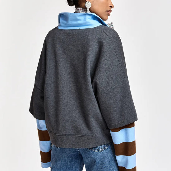 A woman shown from the back wearing a dark gray oversized pullover with a bright blue collar and light blue and brown striped sleeves, paired with blue jeans, against a plain light gray studio background.