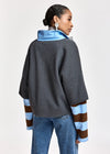 A woman shown from the back wearing a dark gray oversized pullover with a bright blue collar and light blue and brown striped sleeves, paired with blue jeans, against a plain light gray studio background.