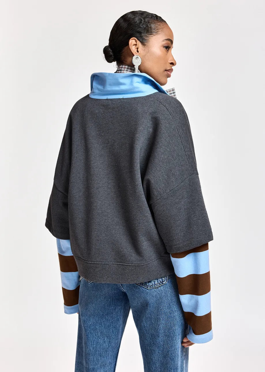 A woman shown from the back wearing a dark gray oversized pullover with a bright blue collar and light blue and brown striped sleeves, paired with blue jeans, against a plain light gray studio background.