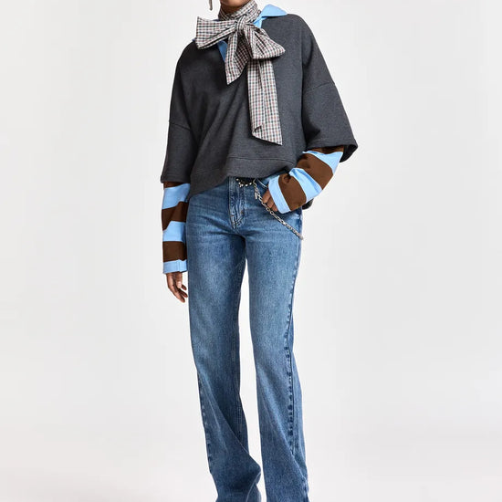 A model stands against a light gray studio background wearing a dark gray oversized top with striped brown and light blue sleeves, blue jeans, and a patterned scarf tied around the neck.