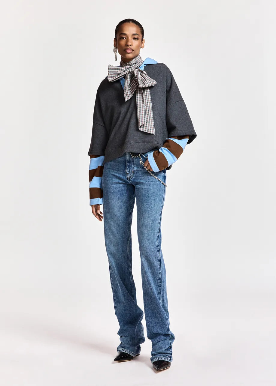 A model stands against a light gray studio background wearing a dark gray oversized top with striped brown and light blue sleeves, blue jeans, and a patterned scarf tied around the neck.
