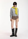 A model stands against a plain white studio background wearing a gray knit sweater, a bright yellow scarf, brown and beige plaid shorts, and black knee-high boots.