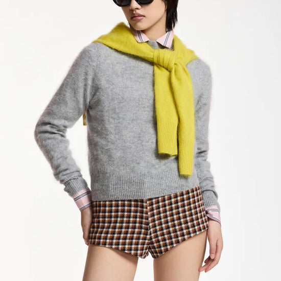 A model wearing a light gray knit sweater, a bright yellow knit scarf, brown and beige plaid shorts, and black sunglasses, posed against a clean white studio background.