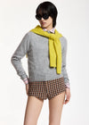A model wearing a light gray knit sweater, a bright yellow knit scarf, brown and beige plaid shorts, and black sunglasses, posed against a clean white studio background.