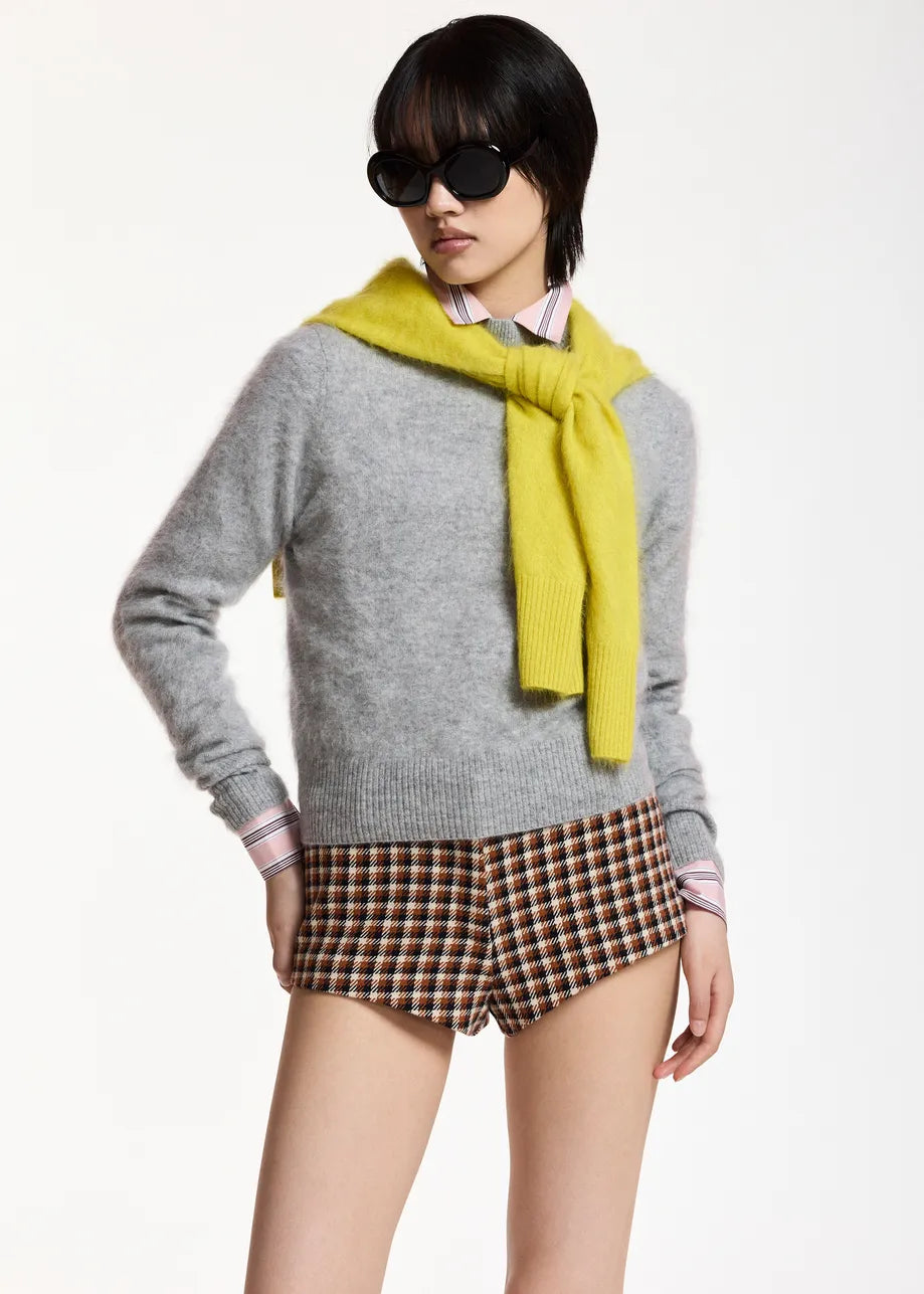 A model wearing a light gray knit sweater, a bright yellow knit scarf, brown and beige plaid shorts, and black sunglasses, posed against a clean white studio background.