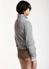 A model shown from the back wearing a light gray knit mock-neck sweater over a pink collared shirt, paired with brown and black plaid shorts, against a plain white background.