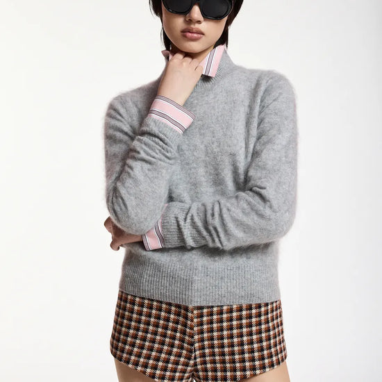 A model wearing a light gray knit sweater with pink and white striped collar and cuffs, paired with brown and beige plaid shorts, against a plain white studio background.