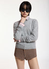 A model wearing a light gray knit sweater with pink and white striped collar and cuffs, paired with brown and beige plaid shorts, against a plain white studio background.