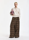 A model stands against a white studio background wearing a cream knit sweater with a tie-neck detail and wide-leg leopard-print trousers. She holds a red, blue, and white plaid clutch.