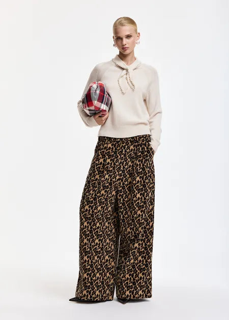A model stands against a white studio background wearing a cream knit sweater with a tie-neck detail and wide-leg leopard-print trousers. She holds a red, blue, and white plaid clutch.