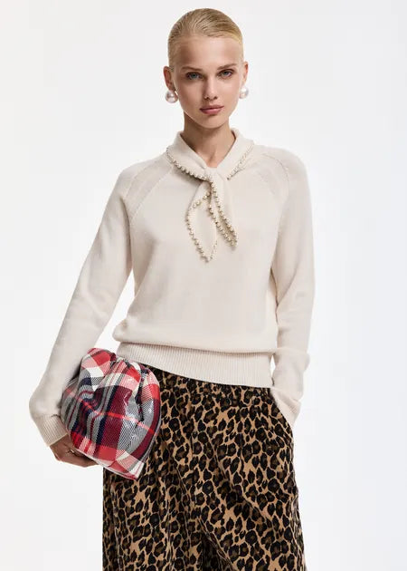 A model wearing a cream knit sweater with a pearl-bead collar and a red plaid clutch bag, paired with a leopard-print skirt, against a plain white studio background.