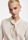 A close-up of a blonde model wearing a cream-colored knit sweater with a pearl-trimmed tie collar and matching pearl drop embellishments.