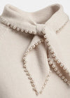 Close-up of a cream-colored ribbed knit top featuring a large bow at the neckline, decorated with strands of white faux pearls.