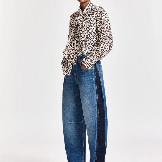 A model stands against a light gray studio background wearing a leopard-print button-up shirt, wide-leg blue jeans with dark side panels, and brown pointed-toe boots.