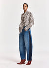 A model stands against a light gray studio background wearing a leopard-print button-up shirt, wide-leg blue jeans with dark side panels, and brown pointed-toe boots.