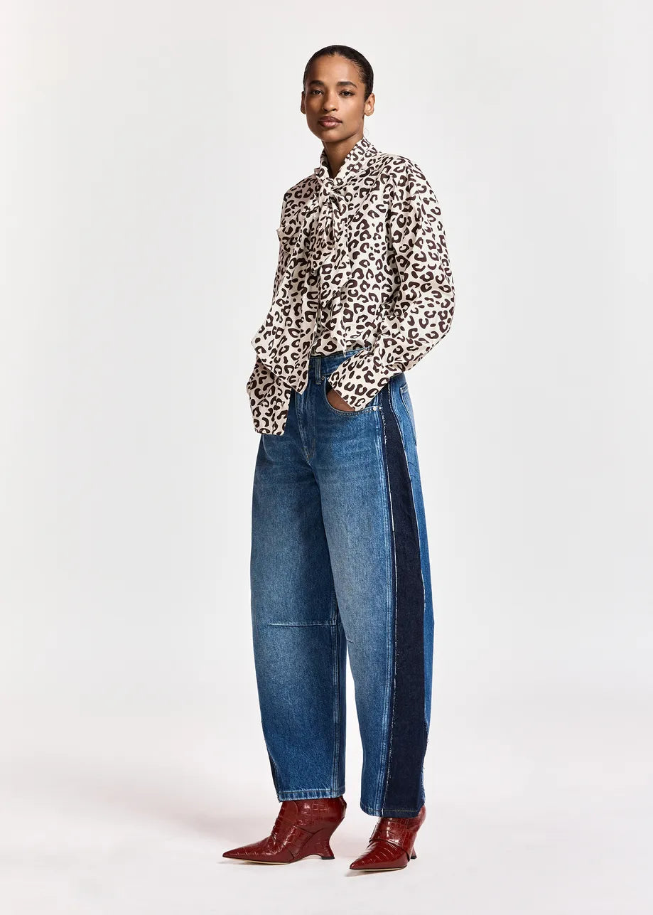 A model stands against a light gray studio background wearing a leopard-print button-up shirt, wide-leg blue jeans with dark side panels, and brown pointed-toe boots.