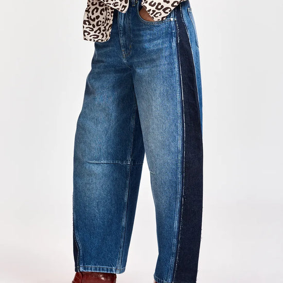 A person wearing wide-leg blue denim jeans with dark navy side panels, paired with a leopard-print top and red pointed-toe heeled boots, shown against a plain light gray studio background.