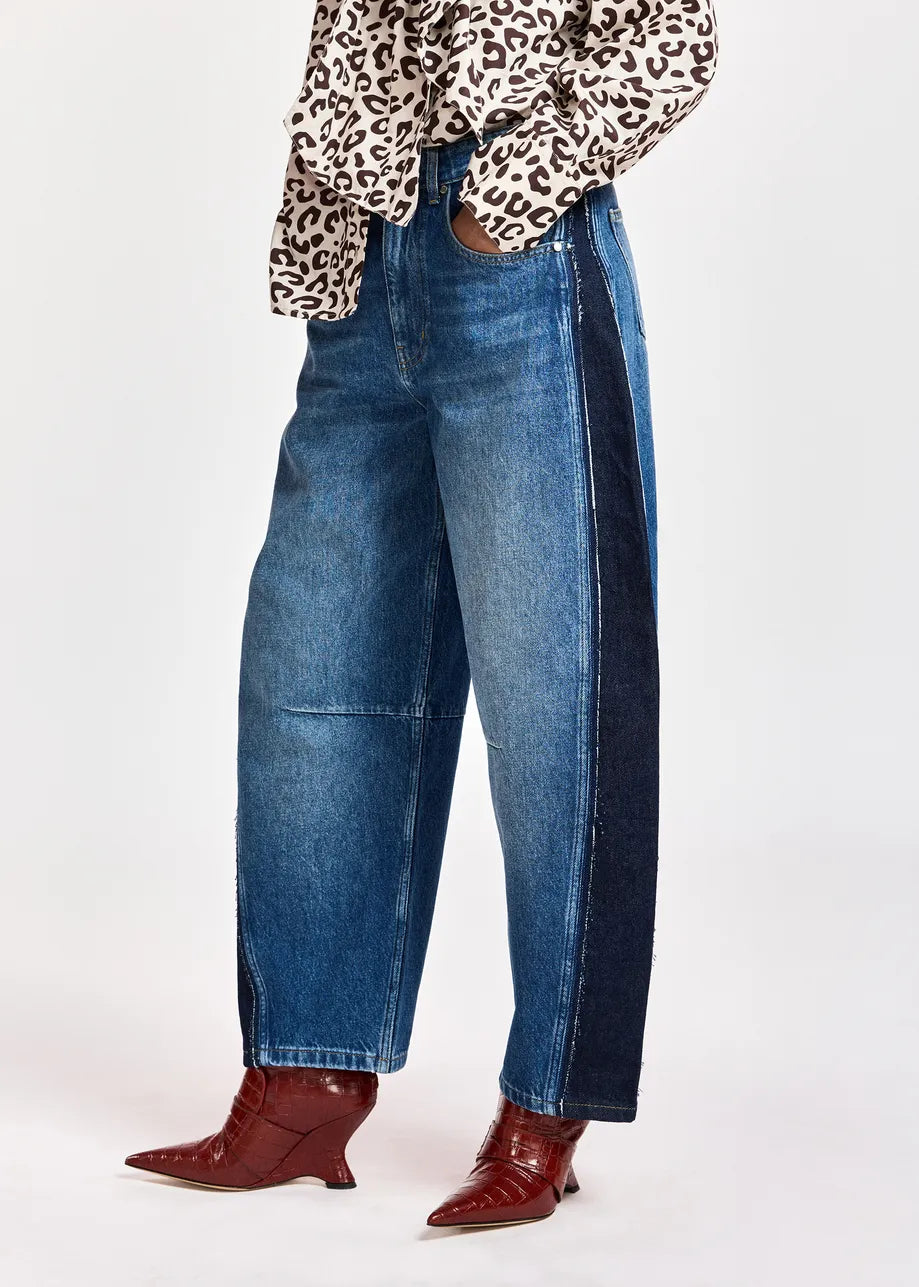 A person wearing wide-leg blue denim jeans with dark navy side panels, paired with a leopard-print top and red pointed-toe heeled boots, shown against a plain light gray studio background.