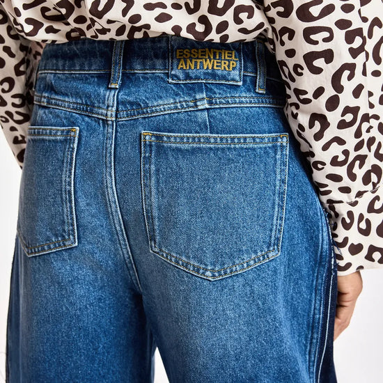 Close-up back view of a person wearing blue denim jeans with a leopard-print long-sleeve top. The waistband shows a gold-tone patch reading 'ESSENTIELS ANTWERP'.