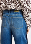 Close-up back view of a person wearing blue denim jeans with a leopard-print long-sleeve top. The waistband shows a gold-tone patch reading 'ESSENTIELS ANTWERP'.