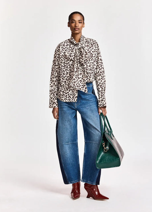 A model stands against a light gray studio background wearing a beige and black leopard-print long-sleeve blouse with a tie-neck detail, wide-leg blue jeans, burgundy pointed-toe heeled boots, and holding a green tote bag.