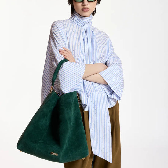 A model wearing a light blue and white striped high-neck shirt, brown wide-leg trousers, green sunglasses, and carrying a large dark green suede shoulder bag against a plain white studio background.