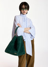 A model wearing a light blue and white striped high-neck shirt, brown wide-leg trousers, green sunglasses, and carrying a large dark green suede shoulder bag against a plain white studio background.