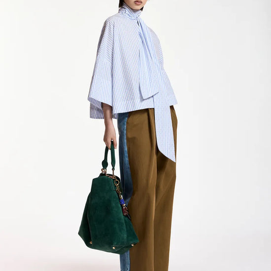 A model stands against a plain white studio background wearing a light blue and white vertically striped high-neck blouse with wide bell sleeves, brown wide-leg trousers, blue jeans partially visible underneath, dark green sunglasses, and holding a dark green suede tote bag.