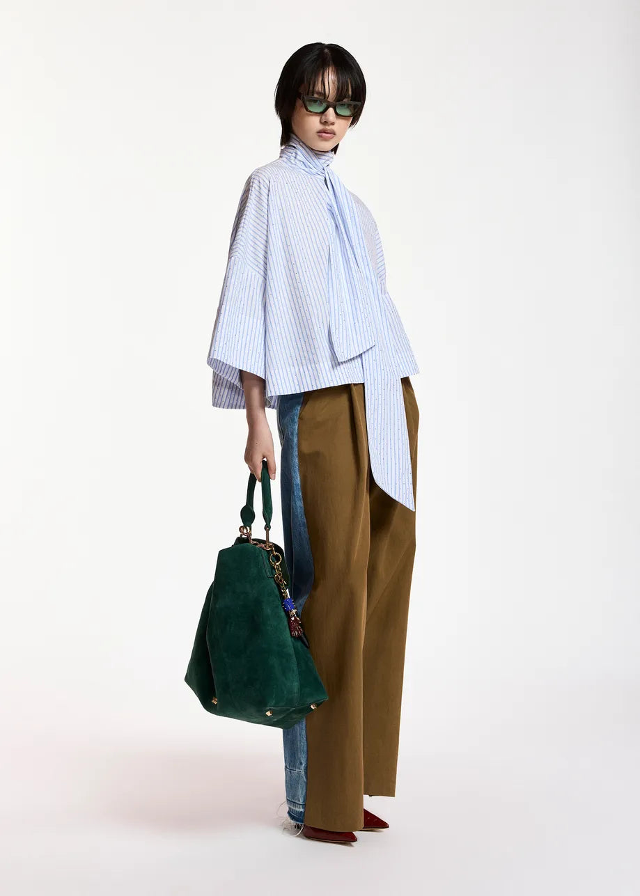 A model stands against a plain white studio background wearing a light blue and white vertically striped high-neck blouse with wide bell sleeves, brown wide-leg trousers, blue jeans partially visible underneath, dark green sunglasses, and holding a dark green suede tote bag.