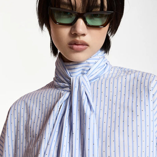 A model with a short black haircut wearing dark brown cat-eye sunglasses and a light blue and white striped shirt with a large bow tie detail at the collar, set against a plain light background.
