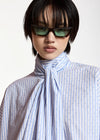 A model with a short black haircut wearing dark brown cat-eye sunglasses and a light blue and white striped shirt with a large bow tie detail at the collar, set against a plain light background.