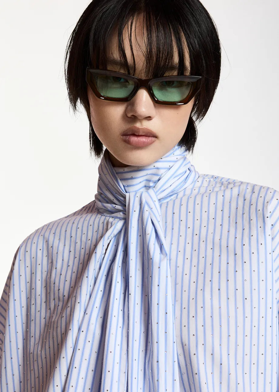 A model with a short black haircut wearing dark brown cat-eye sunglasses and a light blue and white striped shirt with a large bow tie detail at the collar, set against a plain light background.